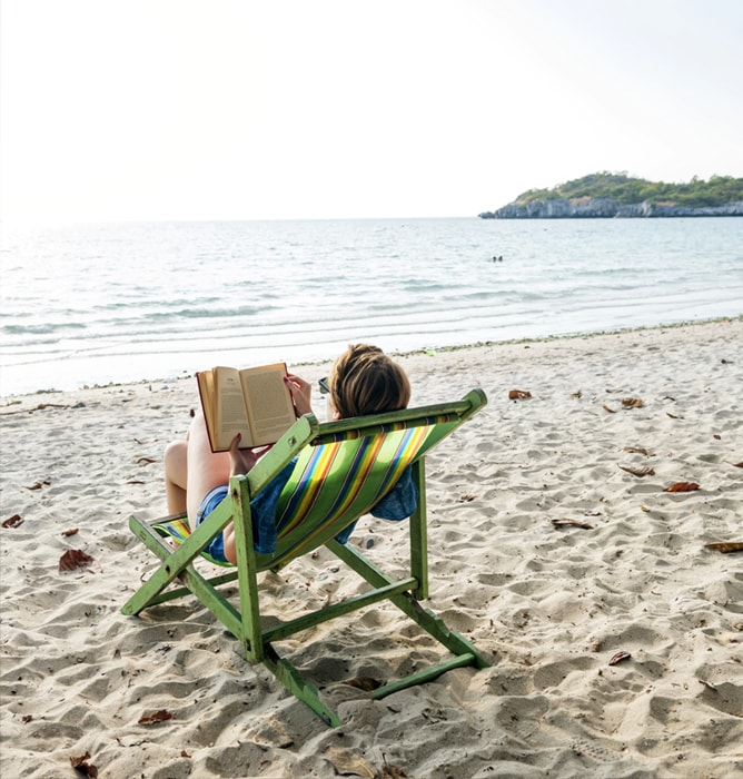 Mulher lendo um livro descansando numa cadeira de praia. Praia com mar calmo e ambiente tranquilo.